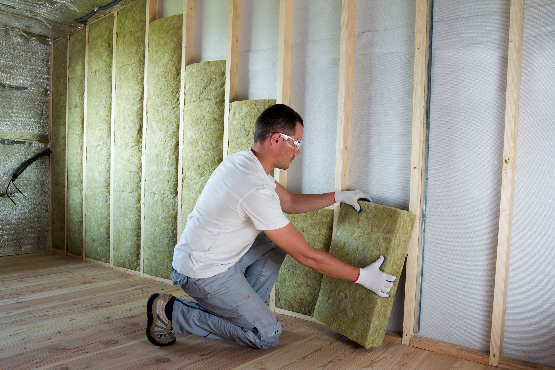 Worker in protective goggles and respirator insulating rock wool insulation in wooden frame for future house walls for cold barrier. Comfortable warm home, economy, construction and renovation concept